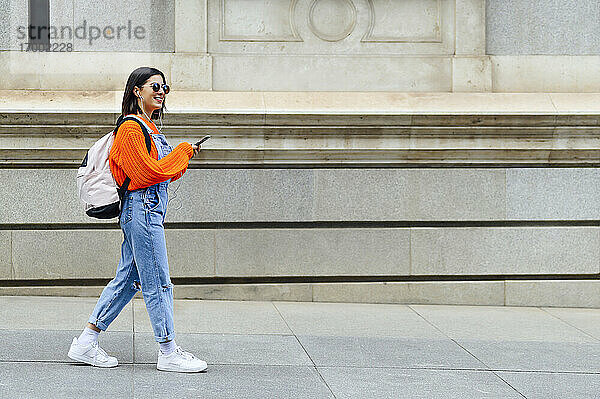 Junge Frau mit Rucksack  die beim Gehen auf dem Fußweg ein Mobiltelefon benutzt
