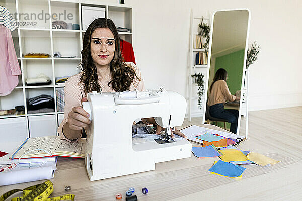 Lächelnde kreative Schneiderin  die in einem Atelier an einer Nähmaschine sitzt