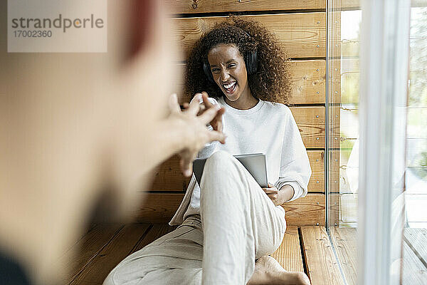 Verspielte Frau mit Kopfhörern und digitalem Tablet  die einem Mann die Hand entgegenstreckt  während sie zu Hause sitzt
