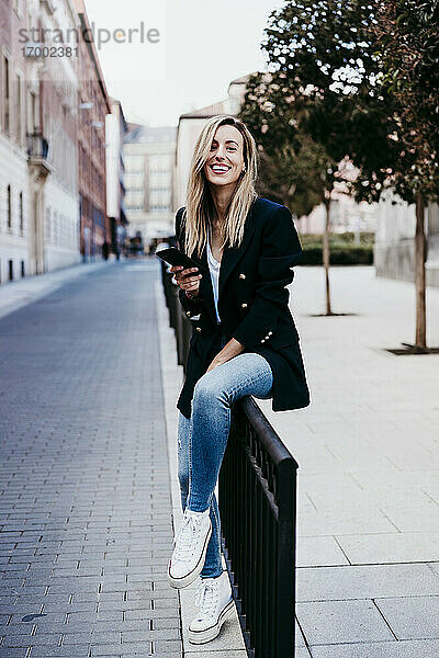 Lächelnde schöne Frau hält Handy beim Sitzen auf Gehweg Zaun in der Stadt