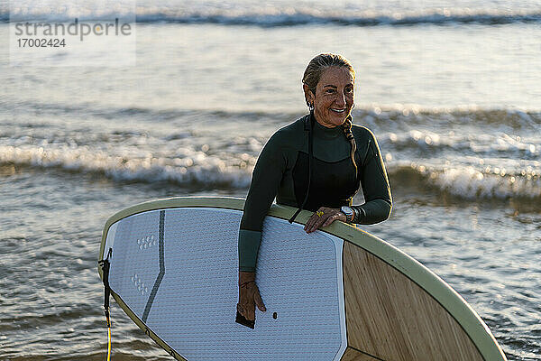 Lächelnde Frau mit Paddleboard im Meer in der Morgendämmerung