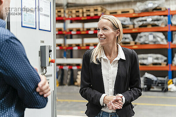 Lächelnde Geschäftsfrau mit verschränkten Händen  die mit einem männlichen Kollegen in einer Fabrik diskutiert