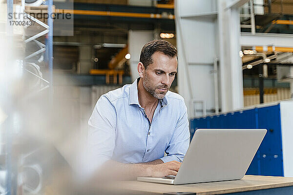 Männlicher Unternehmer arbeitet am Laptop  während er in einer Fabrik sitzt