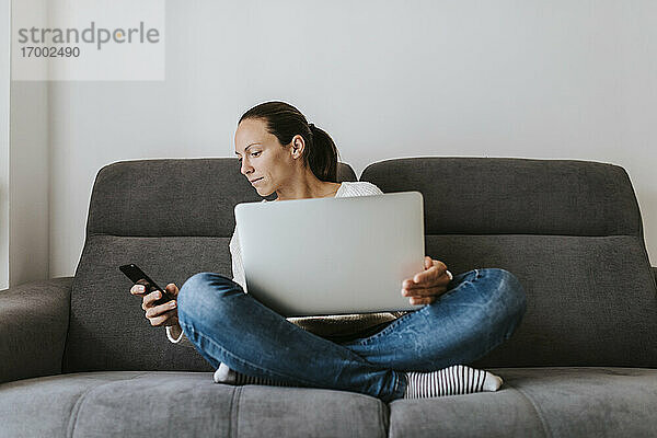 Frau schaut auf ihr Smartphone  während sie mit ihrem Laptop auf dem Sofa sitzt