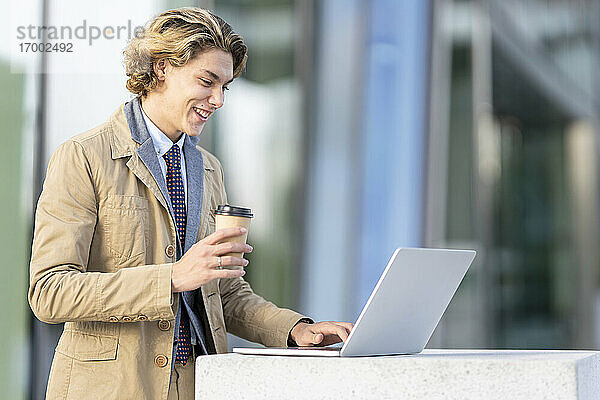 Lächelnder Geschäftsmann mit Kaffeetasse  der im Freien stehend am Laptop arbeitet