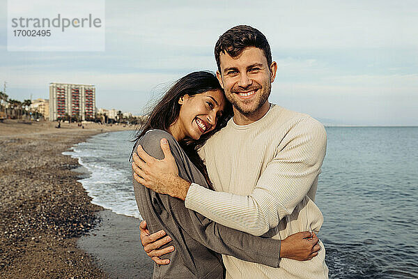 Lächelnde Freundin und Freund umarmen einander gegen den Himmel am Strand