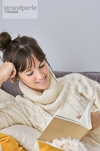 Lächelnde Frau liest ein Buch und entspannt sich auf dem Sofa zu Hause