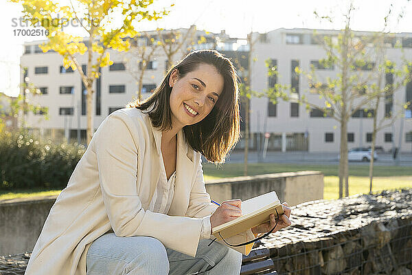 Lächelnde Geschäftsfrau mit Tagebuch auf einer Stützmauer sitzend