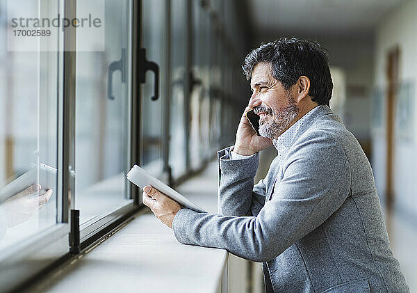Lächelnder männlicher Professor  der am Telefon spricht und durch ein Fenster im Korridor einer Universität schaut