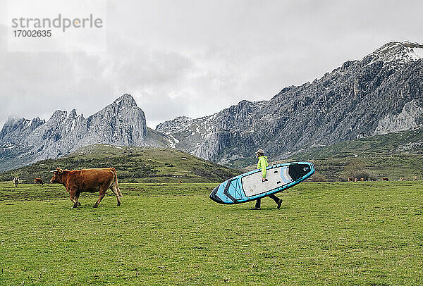 Älterer Mann  der ein Paddelbrett trägt  während er durch eine Landschaft gegen den Himmel läuft  Leon  Spanien