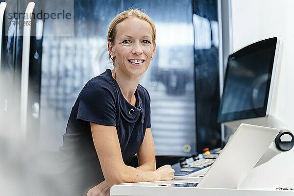 Lächelnde Geschäftsfrau  die sich auf einen Schreibtisch stützt  während sie in der Industrie steht