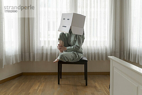Frau mit Karton auf dem Kopf und gelangweiltem Smiley sitzt auf einem Stuhl