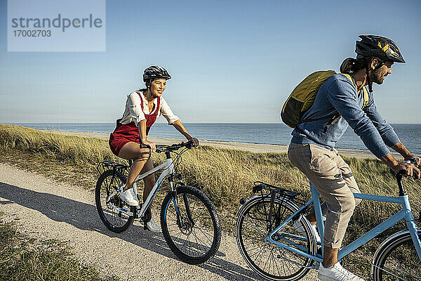Junge Frau lächelt beim Fahrradfahren mit einem Mann auf dem Fußweg gegen das Meer