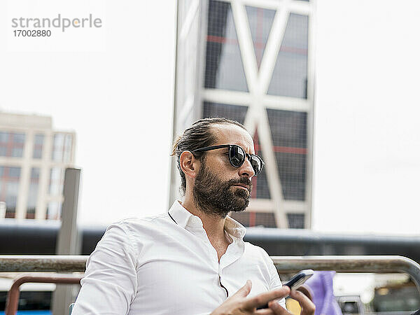Porträt eines bärtigen Geschäftsmannes mit Sonnenbrille  der im Freien sitzt und ein Smartphone in der Hand hält
