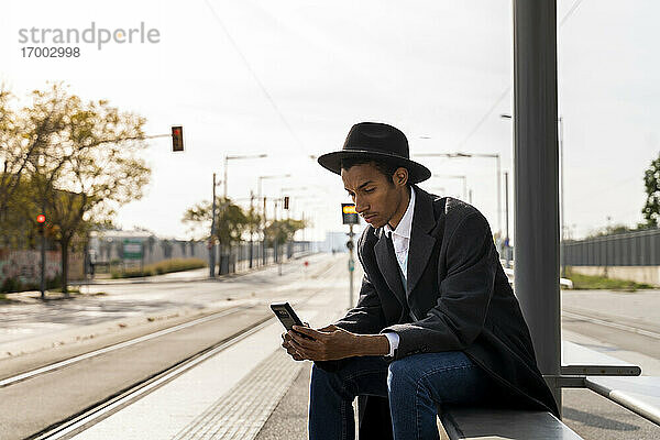 Junger Mann mit Hut benutzt sein Smartphone an einer Bushaltestelle an einem sonnigen Tag