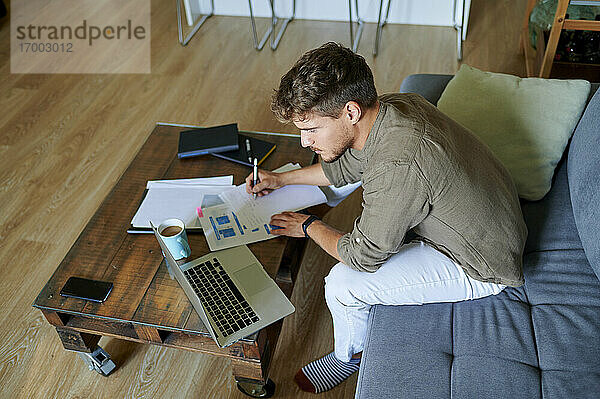 Männlicher Unternehmer  der zu Hause am Laptop sitzt und auf Papier schreibt