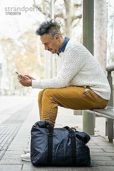 Männlicher Unternehmer  der an der Bushaltestelle sitzt und sein Smartphone benutzt