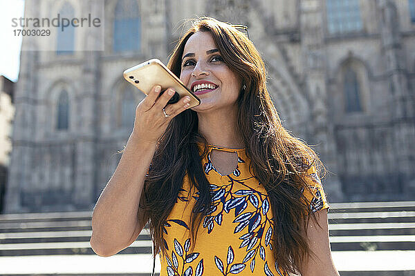 Lächelnde erwachsene Frau  die mit einem Mobiltelefon spricht  während sie am Platz der Kathedrale von Barcelona in Barcelona  Katalonien  Spanien  steht