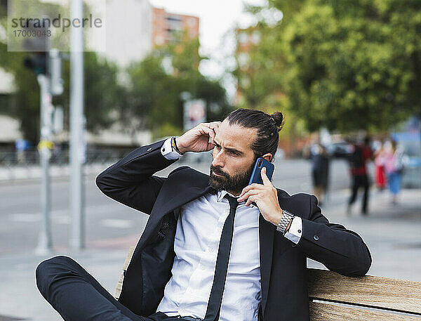 Porträt eines bärtigen Geschäftsmannes  der auf einer Bank sitzt und mit einem Smartphone telefoniert
