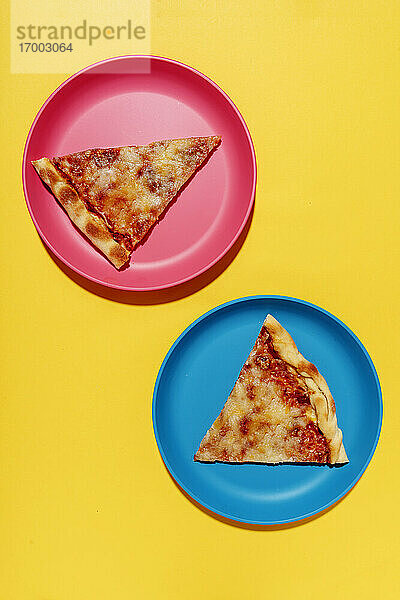 Zwei Scheiben Pizza Margherita auf blauen und rosa Tellern vor gelbem Hintergrund