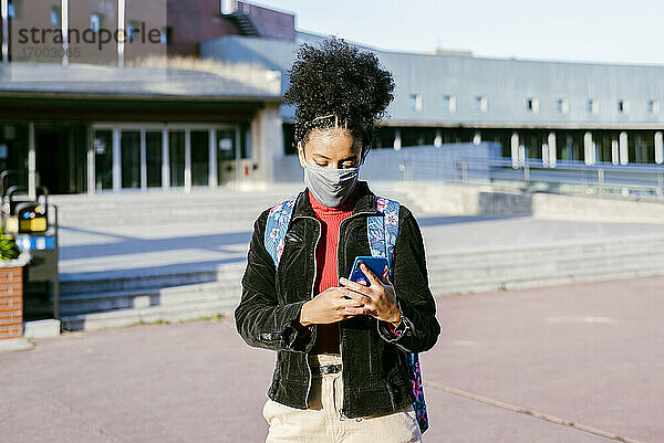 Studentin mit Schutzmaske  die ein Smartphone benutzt  während sie auf der Straße steht