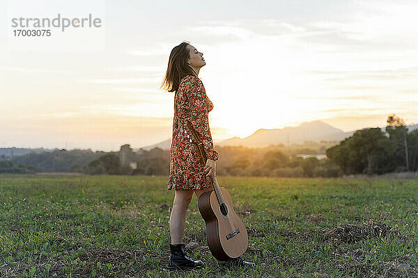 Junge Frau mit geschlossenen Augen hält Gitarre gegen den Himmel bei Sonnenuntergang
