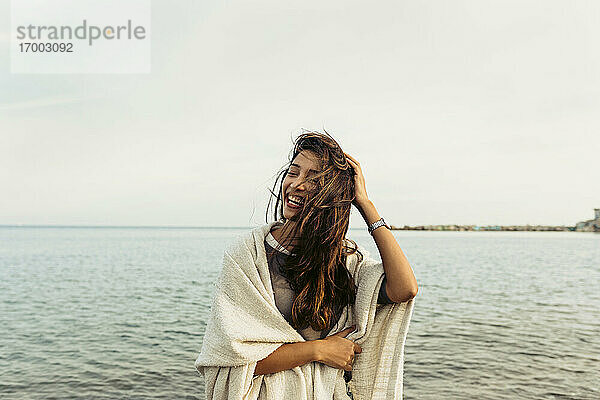 Frau mit Hand in den Haaren  die lachend am Strand am Meer steht
