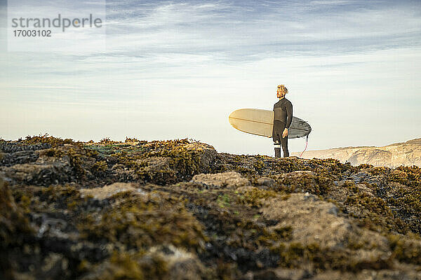 Männlicher Surfer steht mit Surfbrett auf einer Felsformation am Strand gegen den Himmel