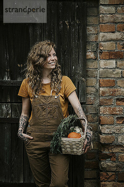Lächelnde Frau hält Gemüsekorb gegen Wand