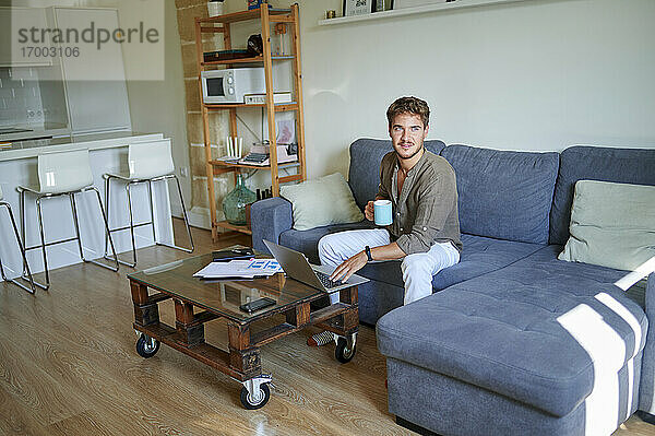 Männlicher Unternehmer hält Kaffeetasse und schaut weg  während er auf dem Sofa im Wohnzimmer sitzt