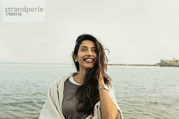Lächelnde Frau mit Hand im Haar  die gegen den klaren Himmel am Strand wegschaut
