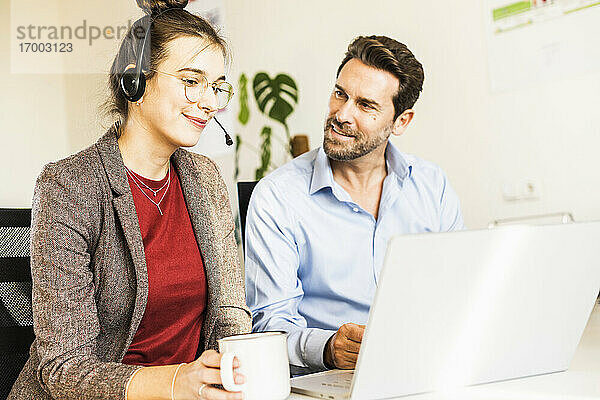 Lächelnde Geschäftsfrau mit Kopfhörern  die Kaffee trinkt  während sie mit einem Kollegen am Laptop im Büro arbeitet
