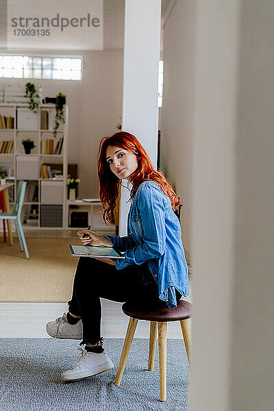 Rothaarige Geschäftsfrau  die ein digitales Tablet benutzt  während sie auf einem Hocker im Büro sitzt