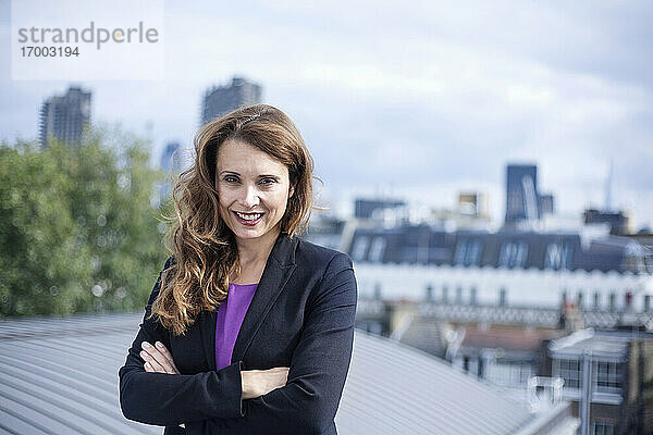 Selbstbewusst lächelnde Unternehmerin  die mit verschränkten Armen auf einer Terrasse vor dem Himmel steht