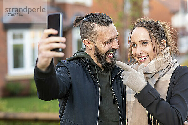 Junge Frau gestikuliert  während sie ein Selfie mit ihrem fröhlichen Freund vor einem neuen Haus macht