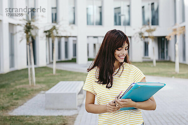 Lächelnde junge Studentin  die ein Buch und einen Ordner auf dem Universitätscampus betrachtet