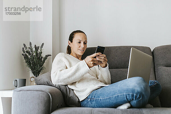 Frau schreibt eine SMS über ihr Smartphone  während sie mit ihrem Laptop auf dem Sofa sitzt