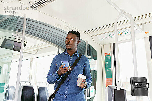Geschäftsmann schaut mit Smartphone und wiederverwendbarem Kaffeebecher im stehenden Zug weg