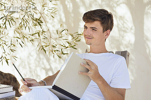 Nachdenklicher junger Mann mit Buch  der im Hinterhof an der Wand sitzt