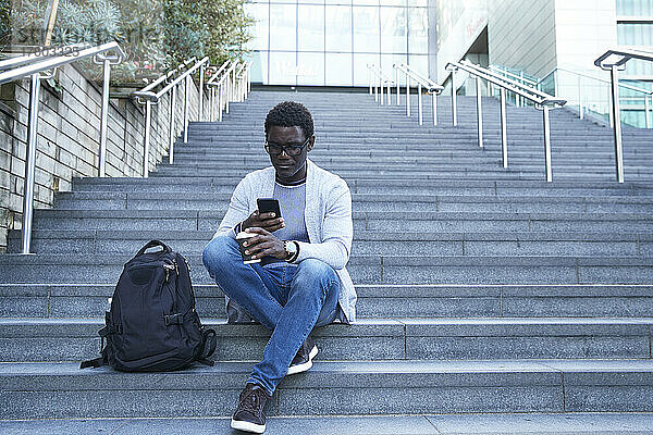Unternehmer  der ein Mobiltelefon benutzt  während er einen Einwegbecher auf einer Treppe im Finanzviertel hält