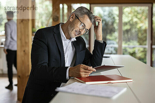 Älterer Geschäftsmann mit Hand im Haar  der ein digitales Tablet benutzt  während er zu Hause am Tisch sitzt