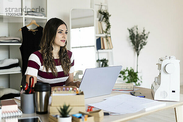 Schöne Modedesignerin schaut weg  während sie mit einem Laptop im Studio sitzt