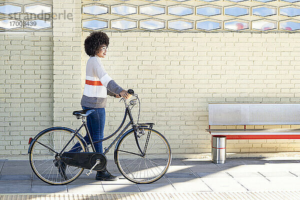 Lächelnde junge Frau  die mit ihrem Fahrrad an einer Backsteinmauer am Bahnhof vorbeifährt