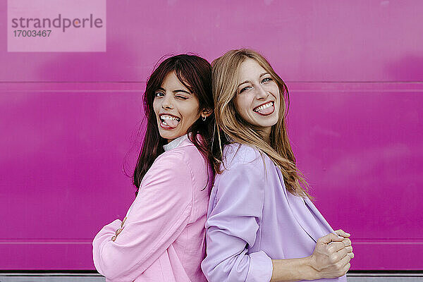 Schwestern strecken die Zunge heraus  während sie Rücken an Rücken an einer rosa Wand stehen