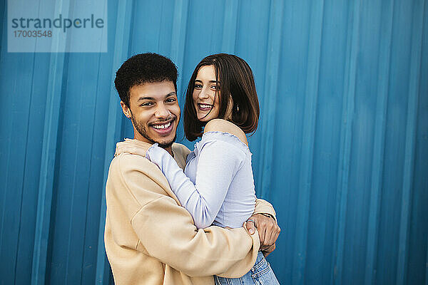 Glückliches junges Paar  das sich gegenseitig umarmt  während es an einer blauen Wand steht