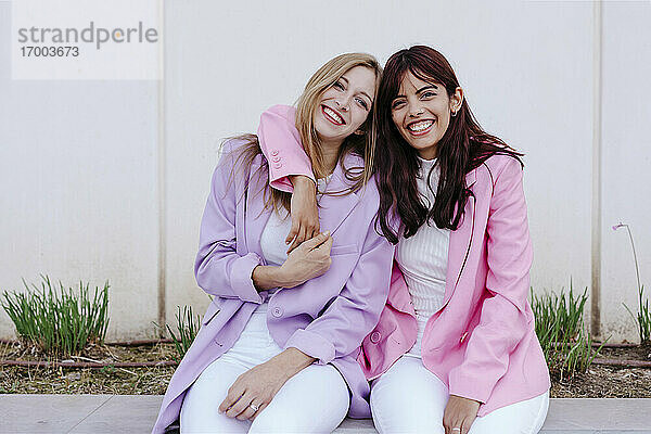 Lächelnde Schwestern mit Arm um sitzen gegen weiße Wand