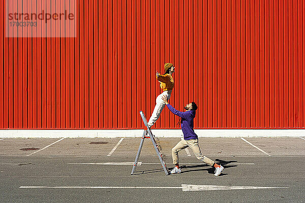 Junger Mann und Frau spielen mit einer Leiter vor einer roten Wand