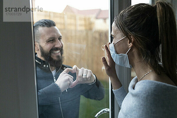 Freund zeigt seiner Freundin durch das Fensterglas eine herzförmige Geste während der COVID-19-Krise