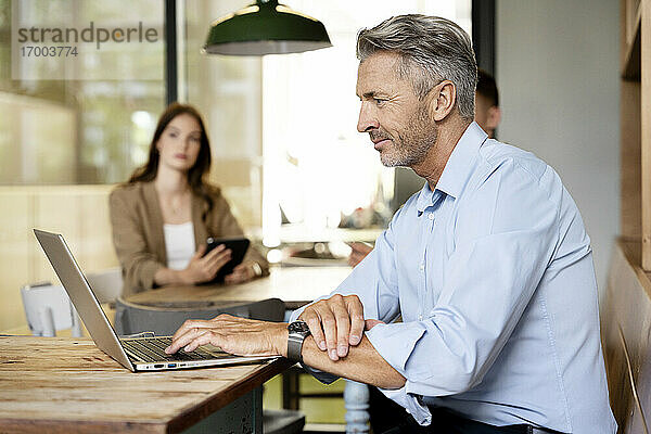 Älterer Geschäftsmann  der an einem Laptop arbeitet  während er mit Kollegen im Hintergrund im Büro sitzt