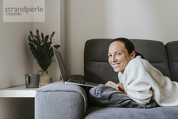 Lächelnde Frau mit Laptop auf dem Sofa im Wohnzimmer liegend
